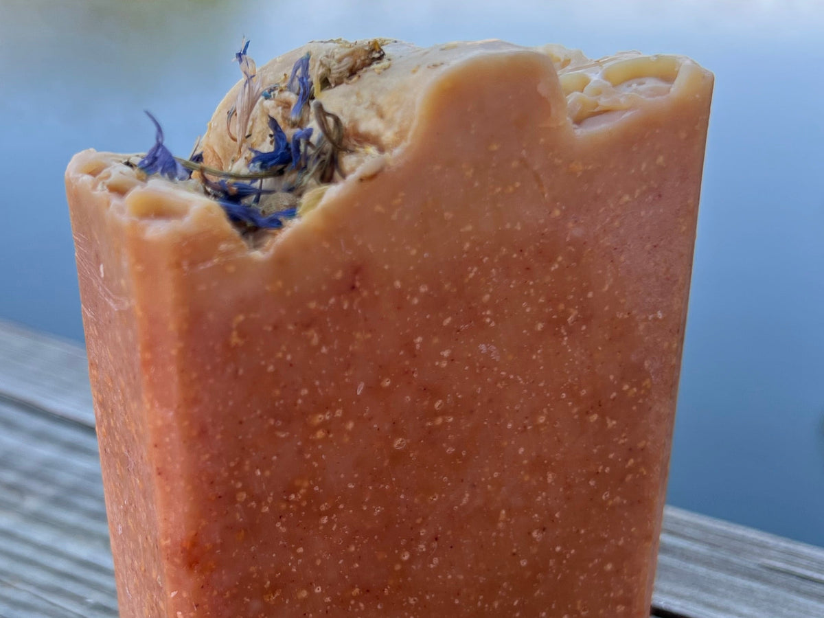 A close-up of a handmade rectangular bar soap in a terracotta orange-brown color, featuring a rustic, rough-branded top edge with dried botanicals, set against a blurred blue background.